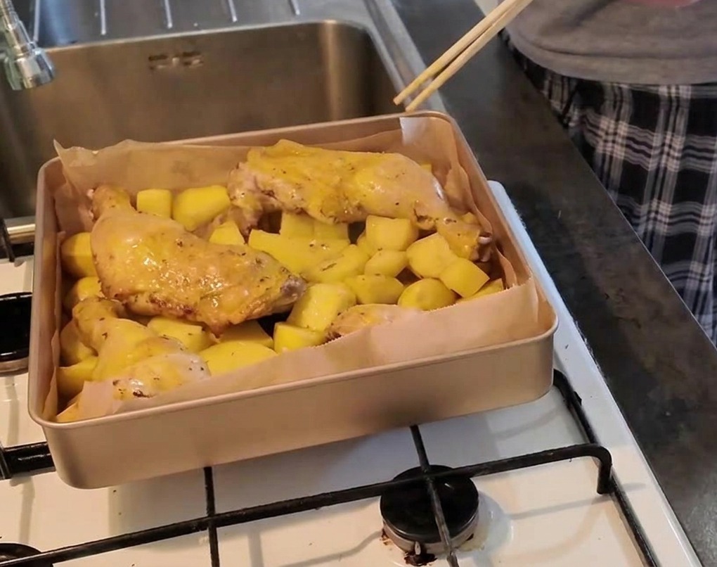 A person uses a pair of chopsticks to poke the meat of a chicken leg resting on a bed of potatoes to verify its doneness.