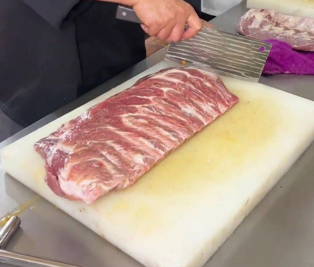 A chef using a large kitchen cleaver to score a diamond pattern onto a raw rack of pork ribs on a white plastic cutting board.
