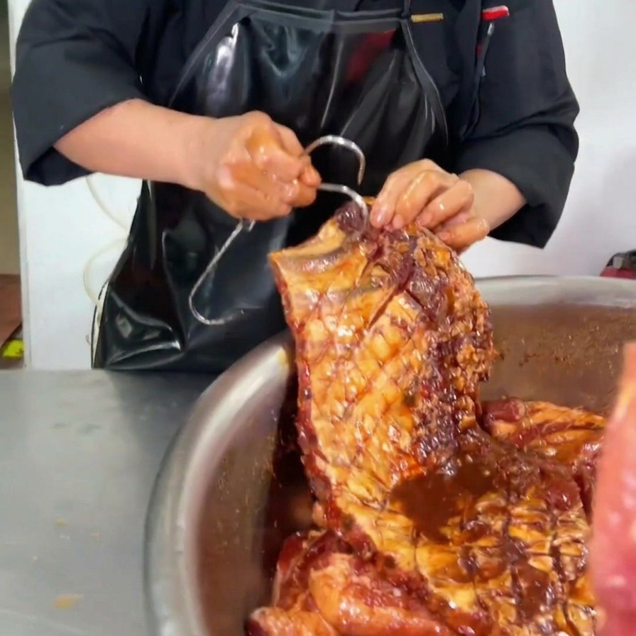 A chef inserting a large metal hook into the upper corner of a fully marinated, dark-sauced rack of pork ribs over a stainless steel bowl.