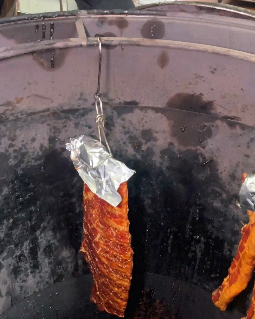 A rack of marinated pork ribs with aluminum foil protecting the top hanging inside a deep, dark cylindrical roasting oven.