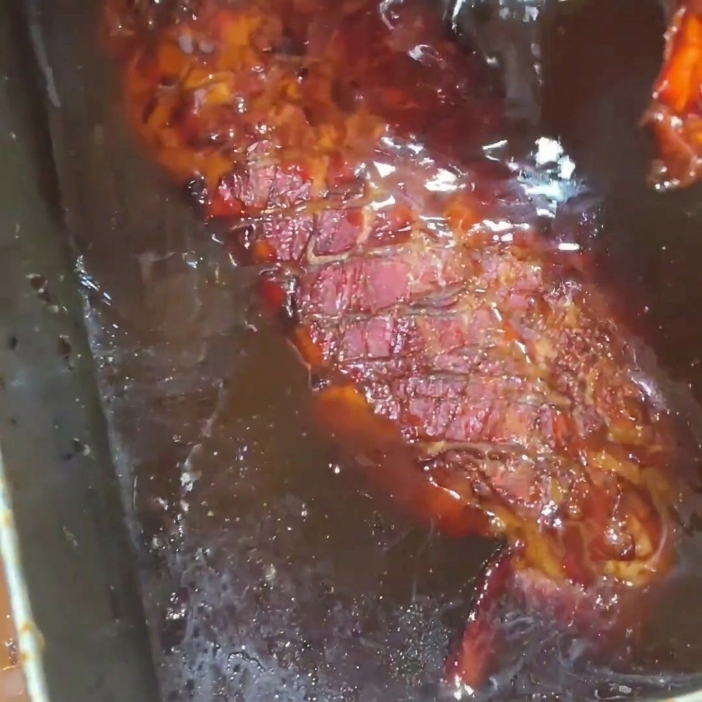 A golden-brown roasted rack of pork ribs being drenched in a rich, glossy honey syrup glaze inside a stainless steel tray.
