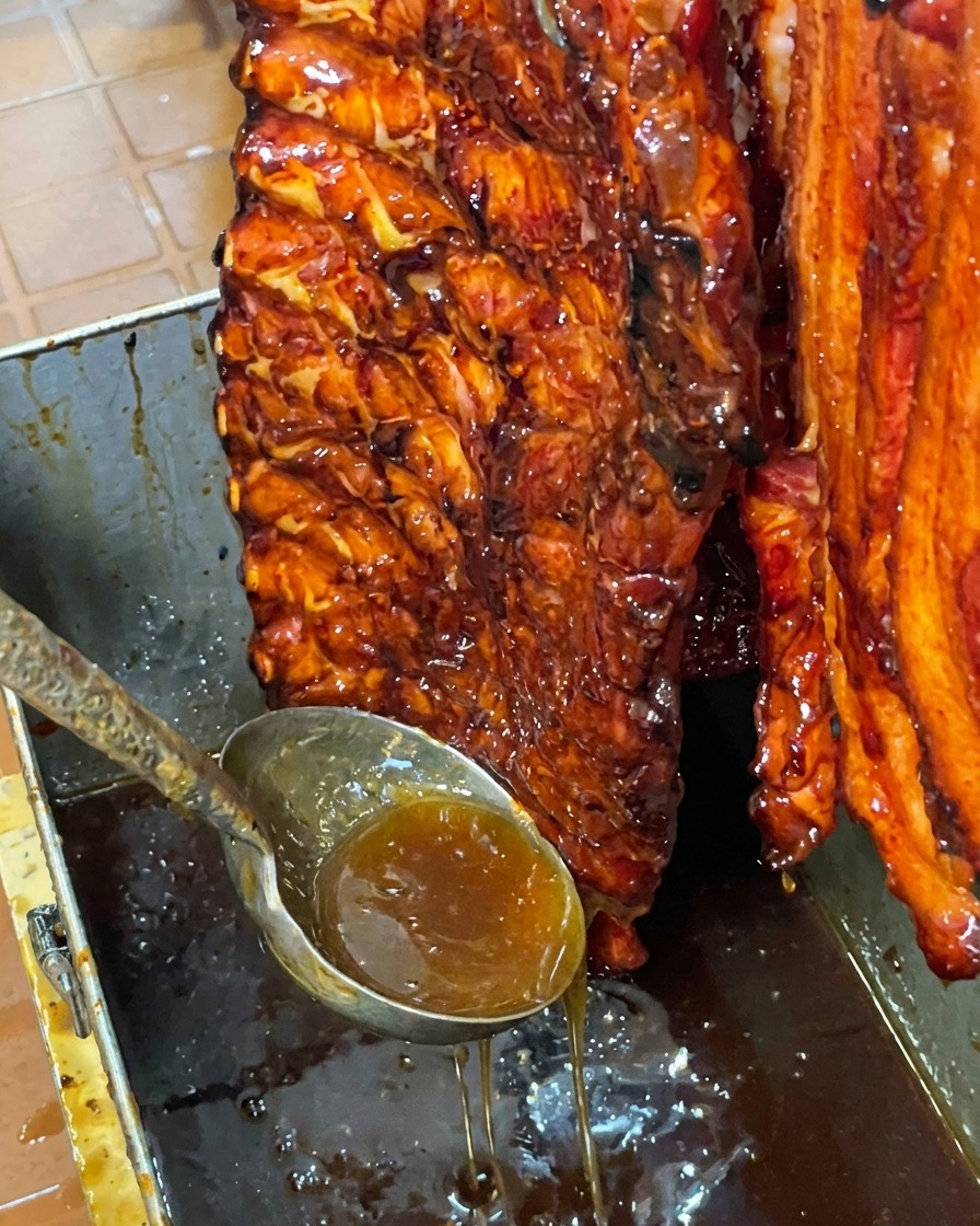 A glistening, honey-glazed rack of pork ribs hanging vertically over a metal basin as excess sweet glaze drips down.