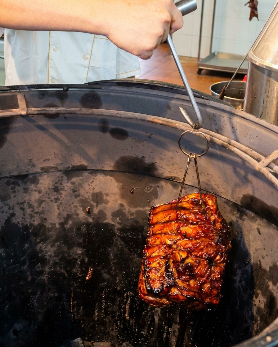 A slab of glistening pork ribs being lowered with a metal hook into a deep, dark cylindrical roasting oven.