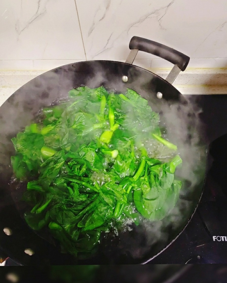 Fresh green leafy vegetables submerged and blanching in a large wok filled with boiling, steaming water.
