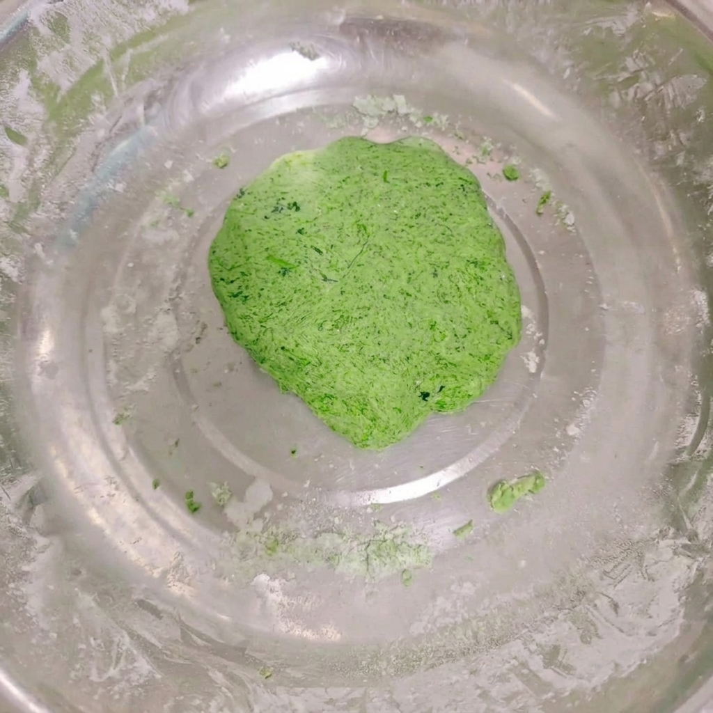 A smooth, vibrant green vegetable dough ball resting inside a large stainless steel mixing bowl with bits of flour around the edges.