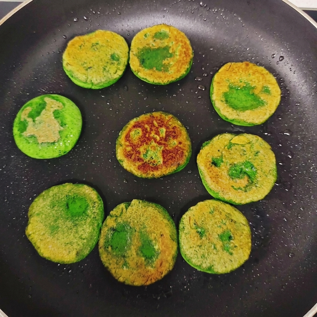 Green vegetable pancakes flipped over in a frying pan, showing uneven golden-brown seared crusts on their tops.