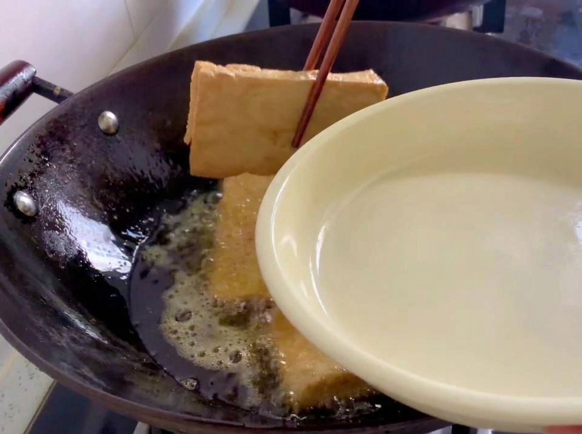 Fried golden-brown tofu slabs being lifted out of a wok with wooden chopsticks.