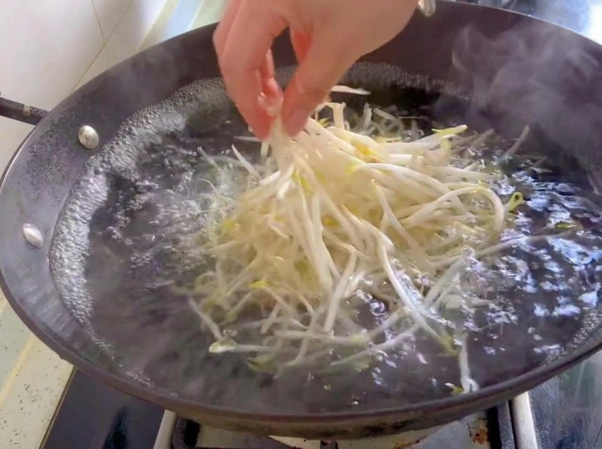 A handful of fresh white bean sprouts being dropped into a wok of boiling water.