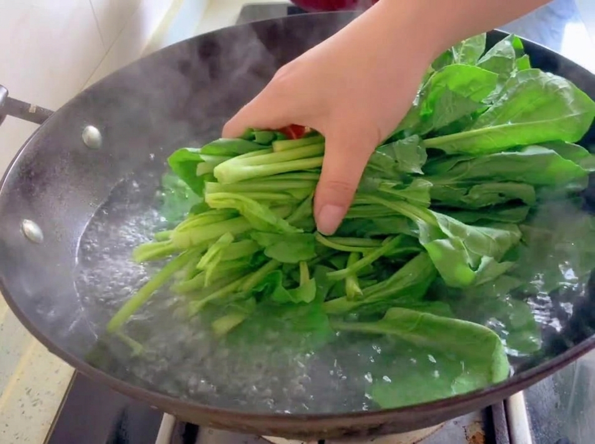 A bundle of fresh green spinach leaves being placed into boiling water in a wok.