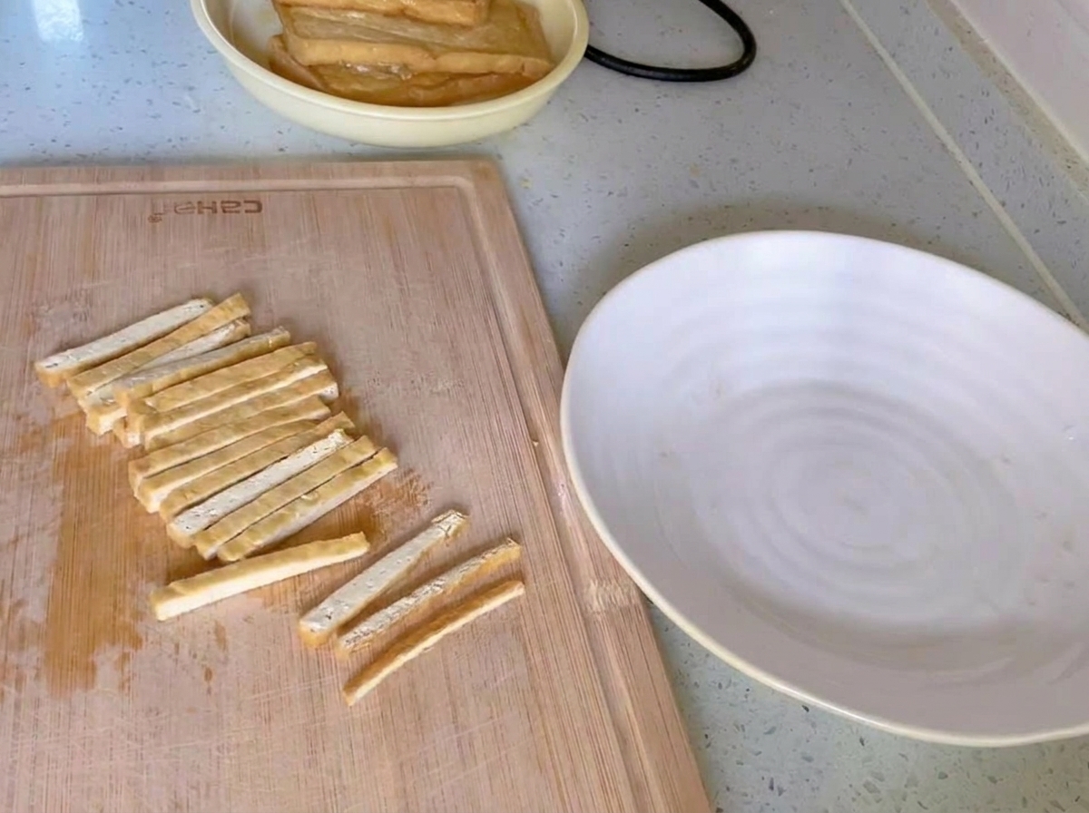 Golden-brown fried tofu sliced into uniform thin strips on a wooden cutting board next to a white plate.