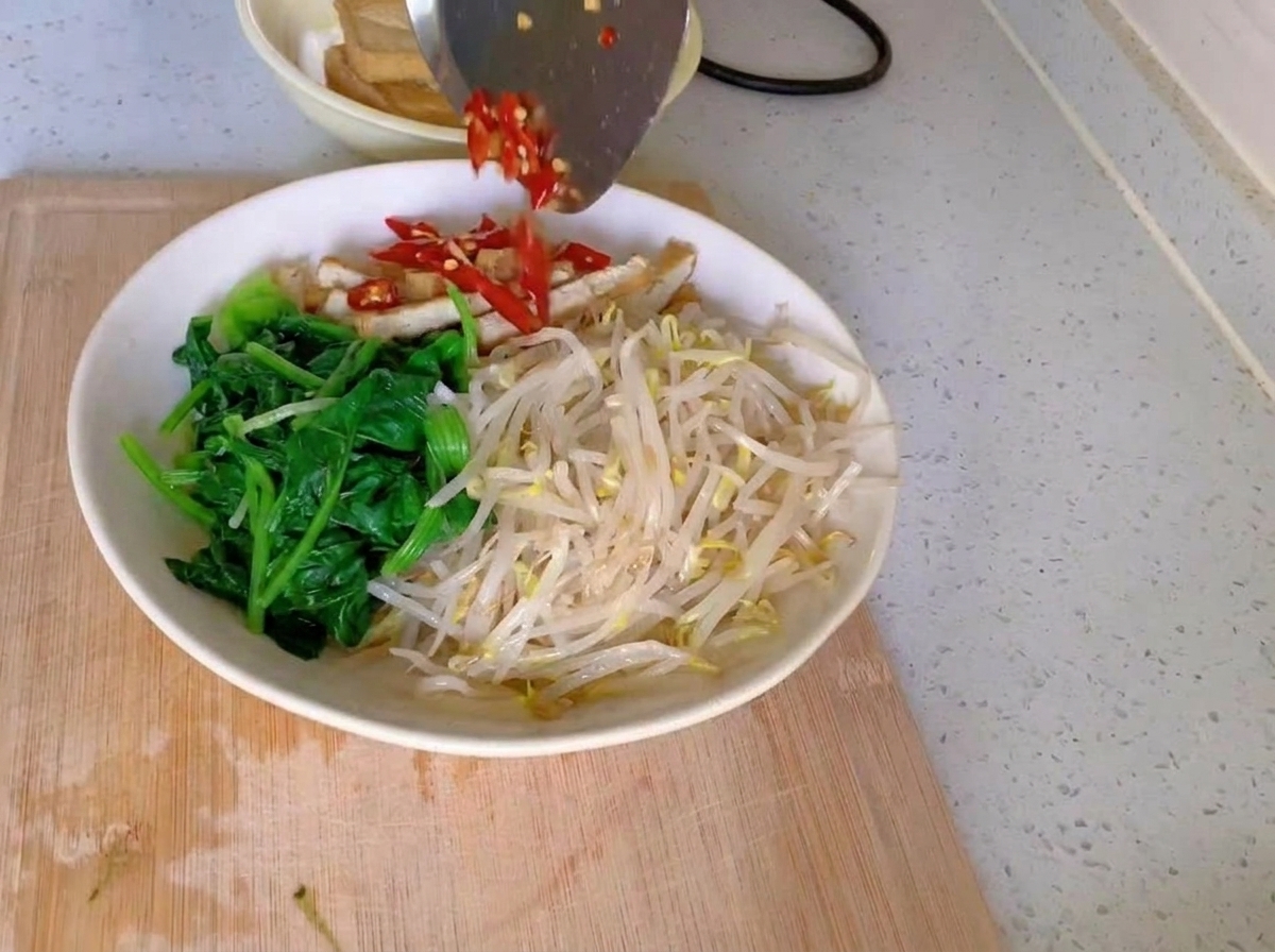 A white bowl filled with distinct sections of vibrant green spinach, pale bean sprouts, and sliced tofu strips, topped with a pile of chopped red chilies and minced garlic.