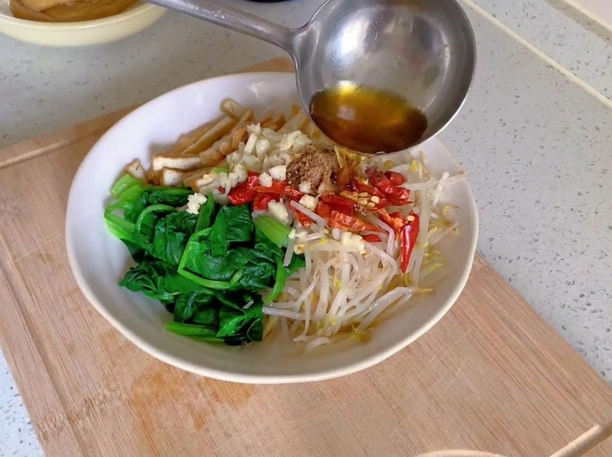 Smoking hot oil being poured from a metal ladle onto minced garlic and bright red dried chilies layered over blanched vegetables in a white bowl.