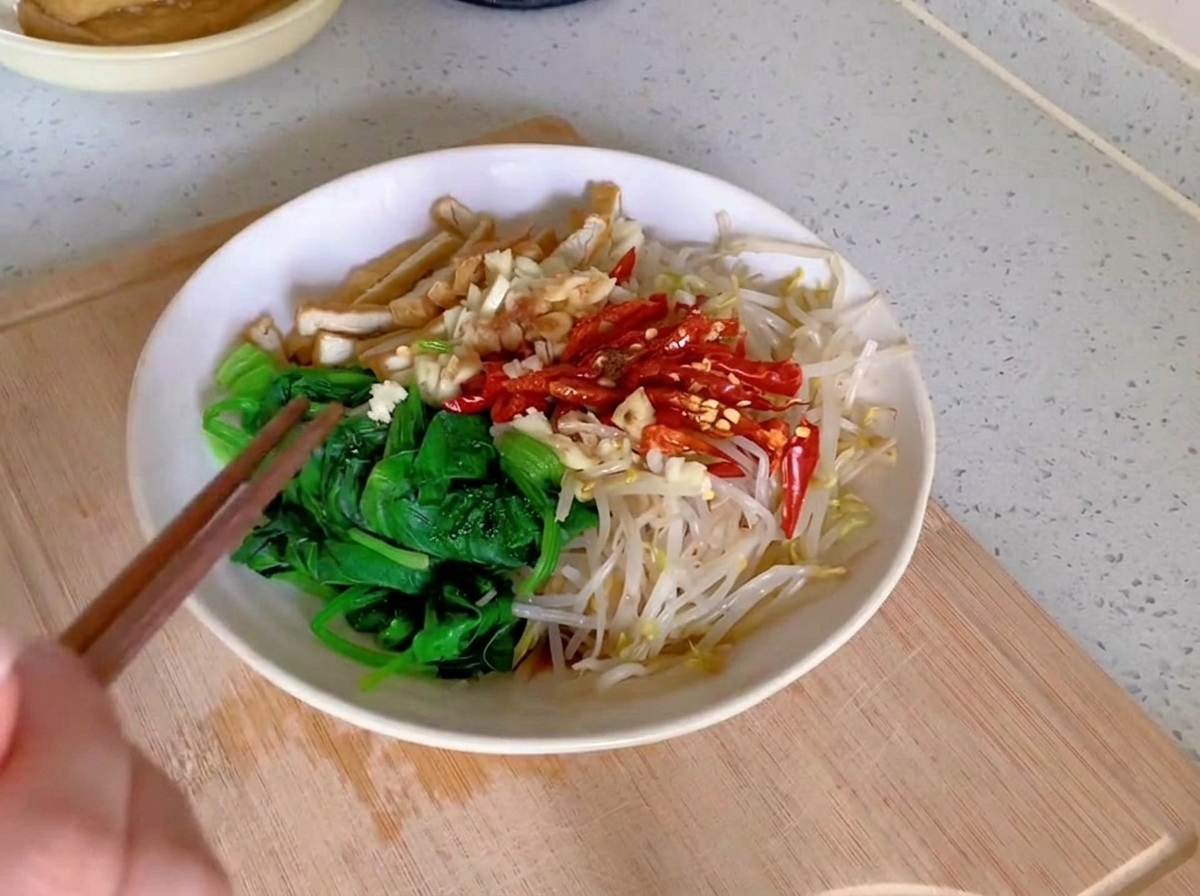 A pair of wooden chopsticks tossing a mixture of green spinach, pale bean sprouts, tofu strips, and red chilies in a white ceramic bowl.