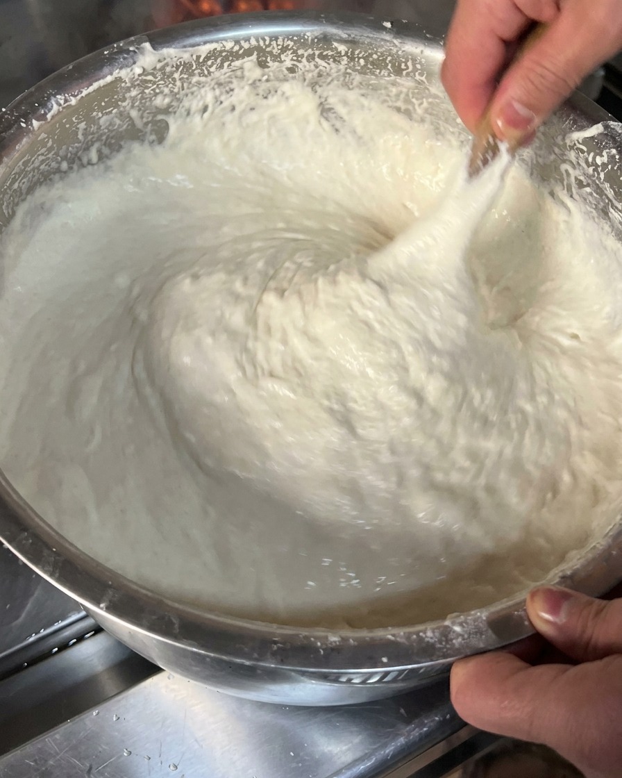 A close-up view of thick, sticky white dough being lifted and stretched with a wooden tool inside a metal mixing bowl.