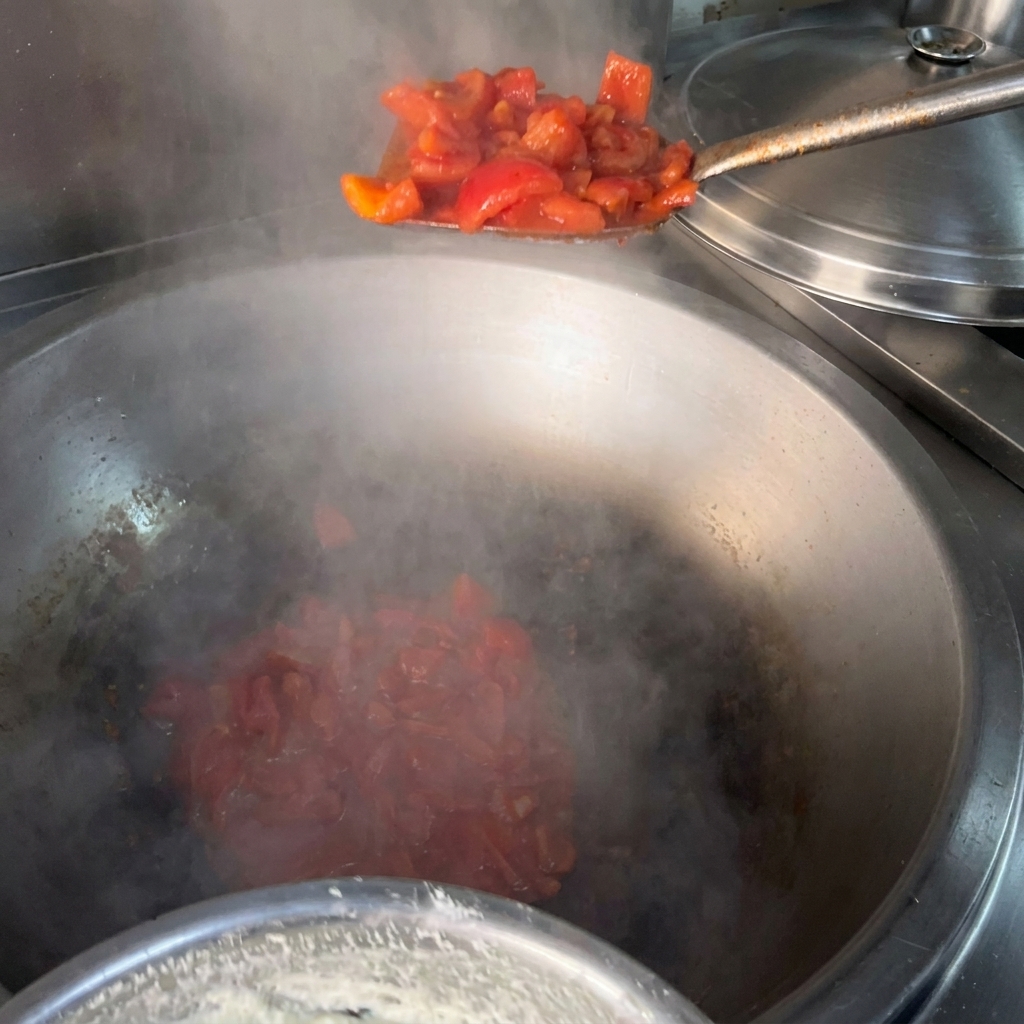 A metal spatula adding chopped red tomatoes into a large, steaming metal wok with hot oil.