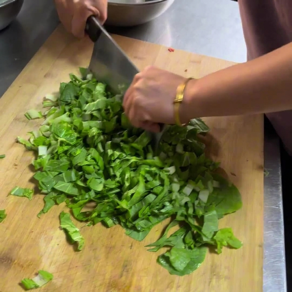 Hands using a large metal cleaver to chop fresh green leafy vegetables on a wooden cutting board.