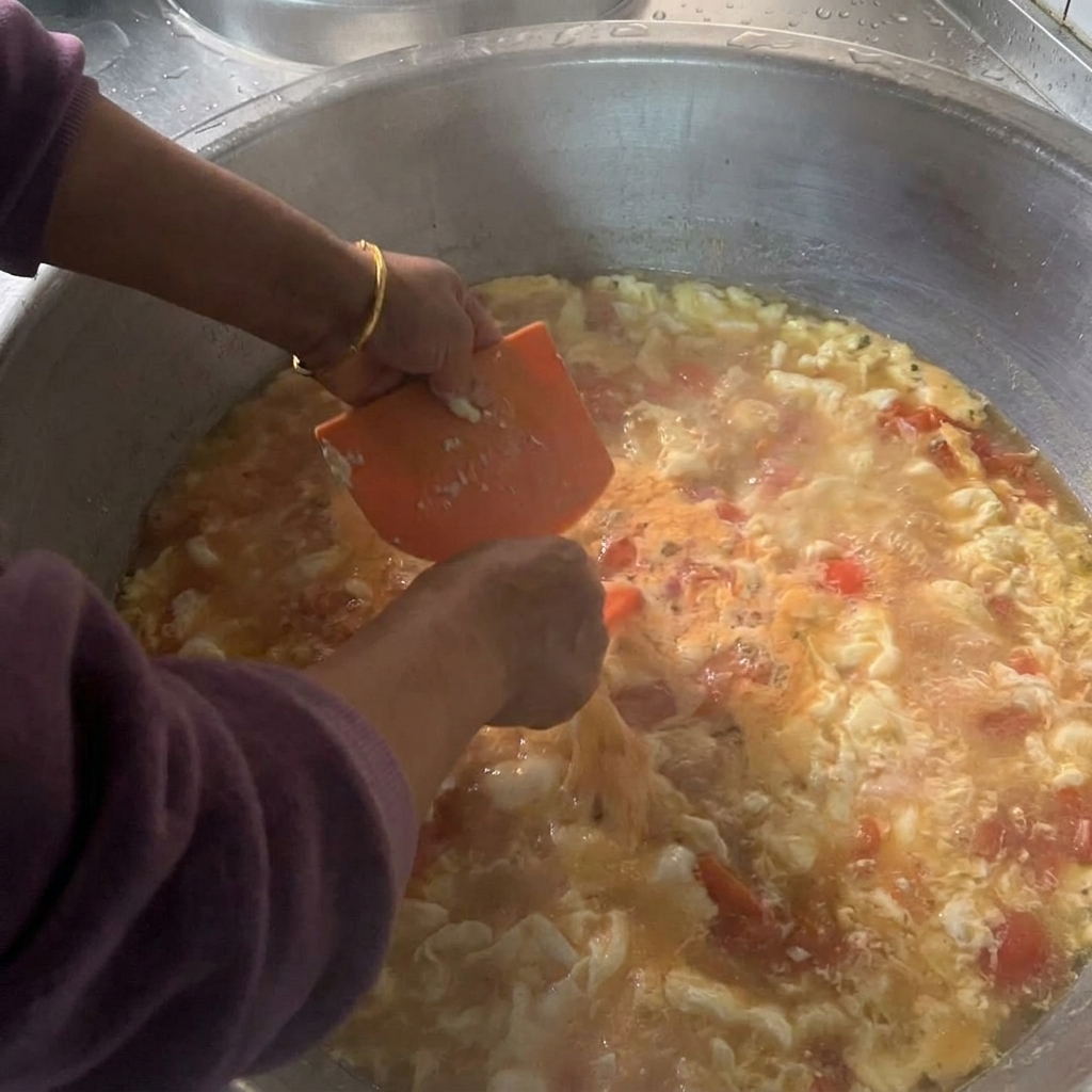 Hands holding an orange plastic scraper to drop small pieces of wet dough into a large pot filled with boiling tomato and egg soup.