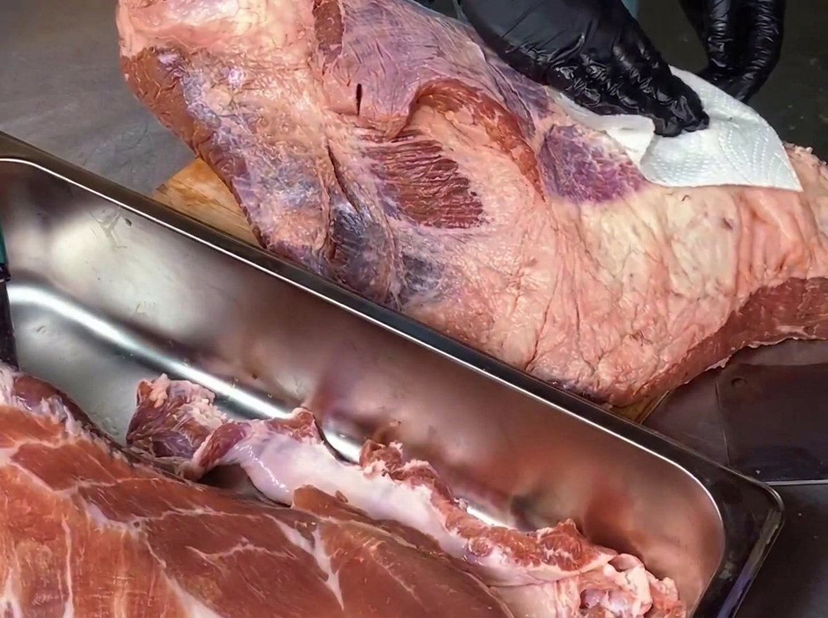 A chef wearing black gloves uses a white paper towel to wipe down a large piece of raw beef brisket on a wooden cutting board, with a stainless steel tray holding trimmings visible in the foreground.