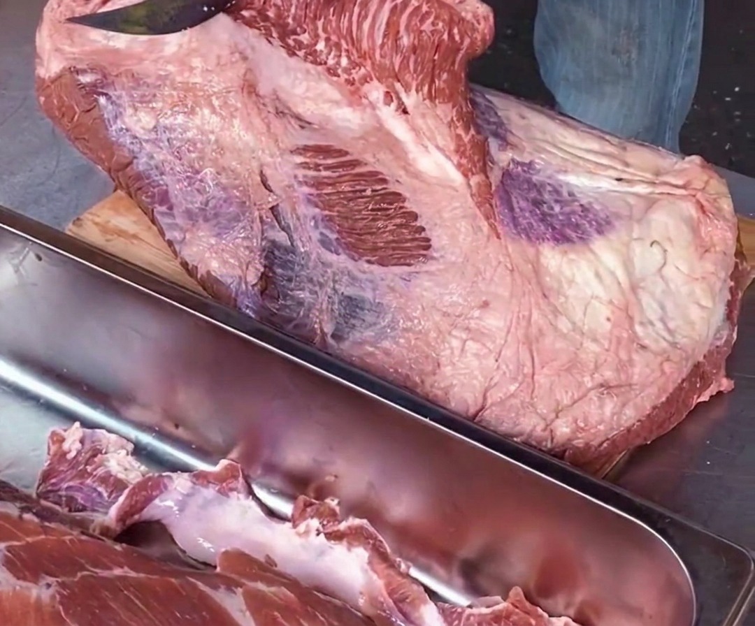 A close-up of a chef using a sharp knife parallel to a cutting board to separate raw point meat from flat meat along a white seam of fat.
