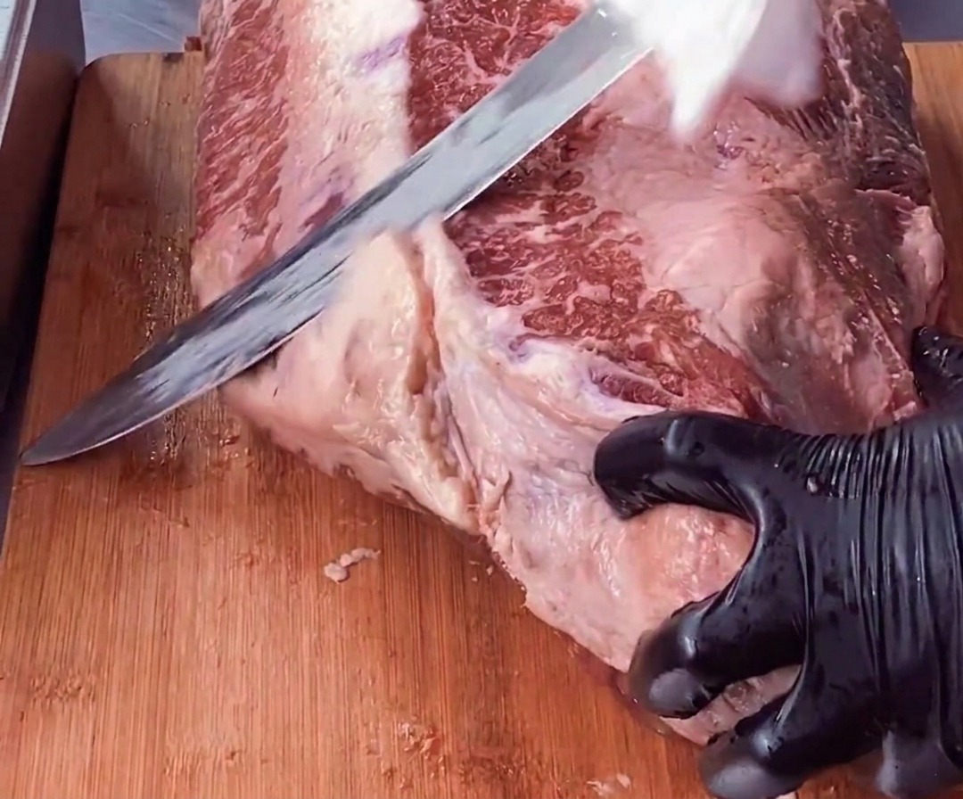 A gloved hand stabilizing a piece of brisket while a long knife trims away a thick layer of white fat from the red meat.