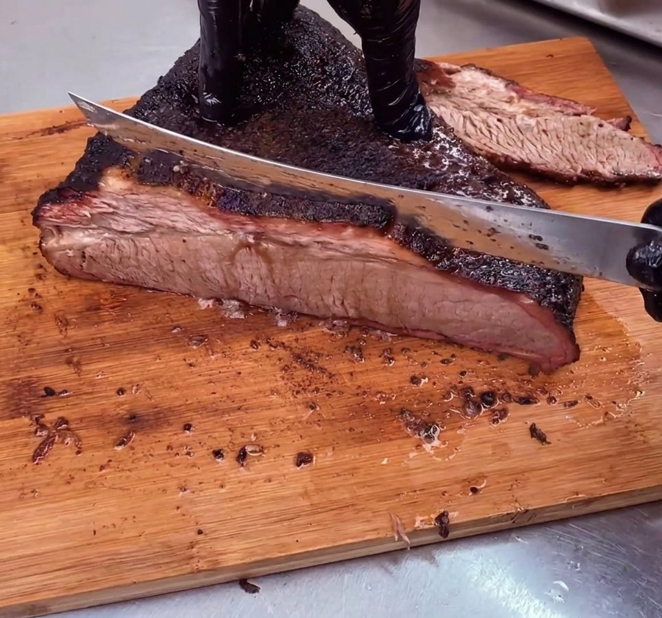 A chef slicing a beautifully smoked beef brisket with a dark bark against the grain into thin slices on a wooden cutting board.