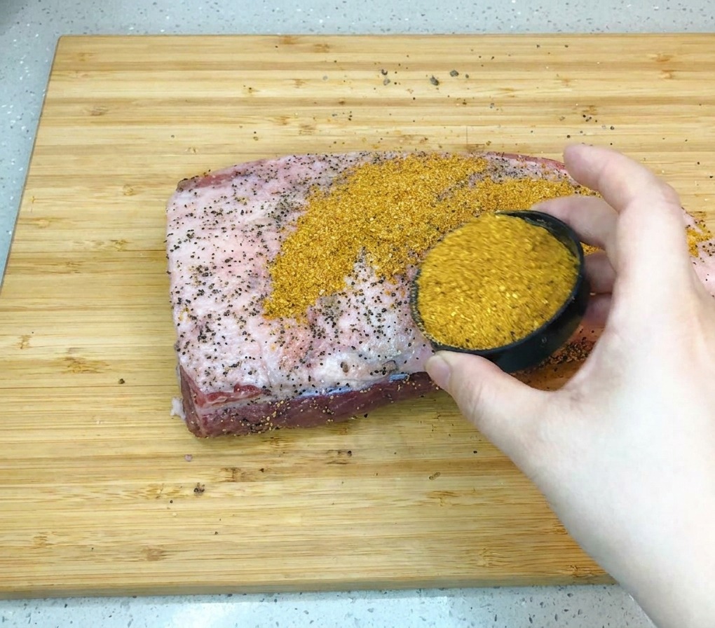 A hand holding a small black bowl and pouring golden-brown barbecue seasoning rub onto a large piece of raw beef brisket on a wooden board.