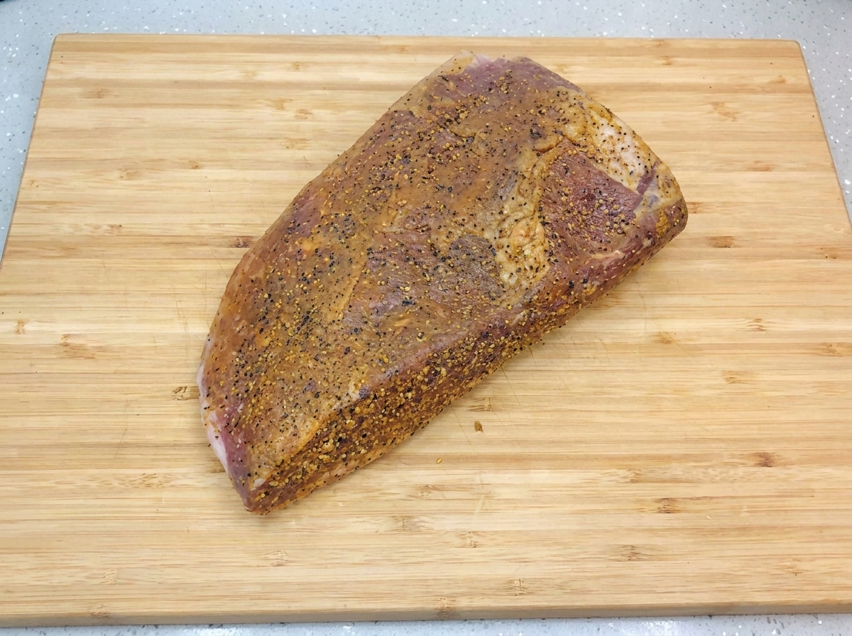 A seasoned, marinated brisket resting on a light-colored wooden cutting board, glistering with a fresh coat of oil and finely ground black pepper.