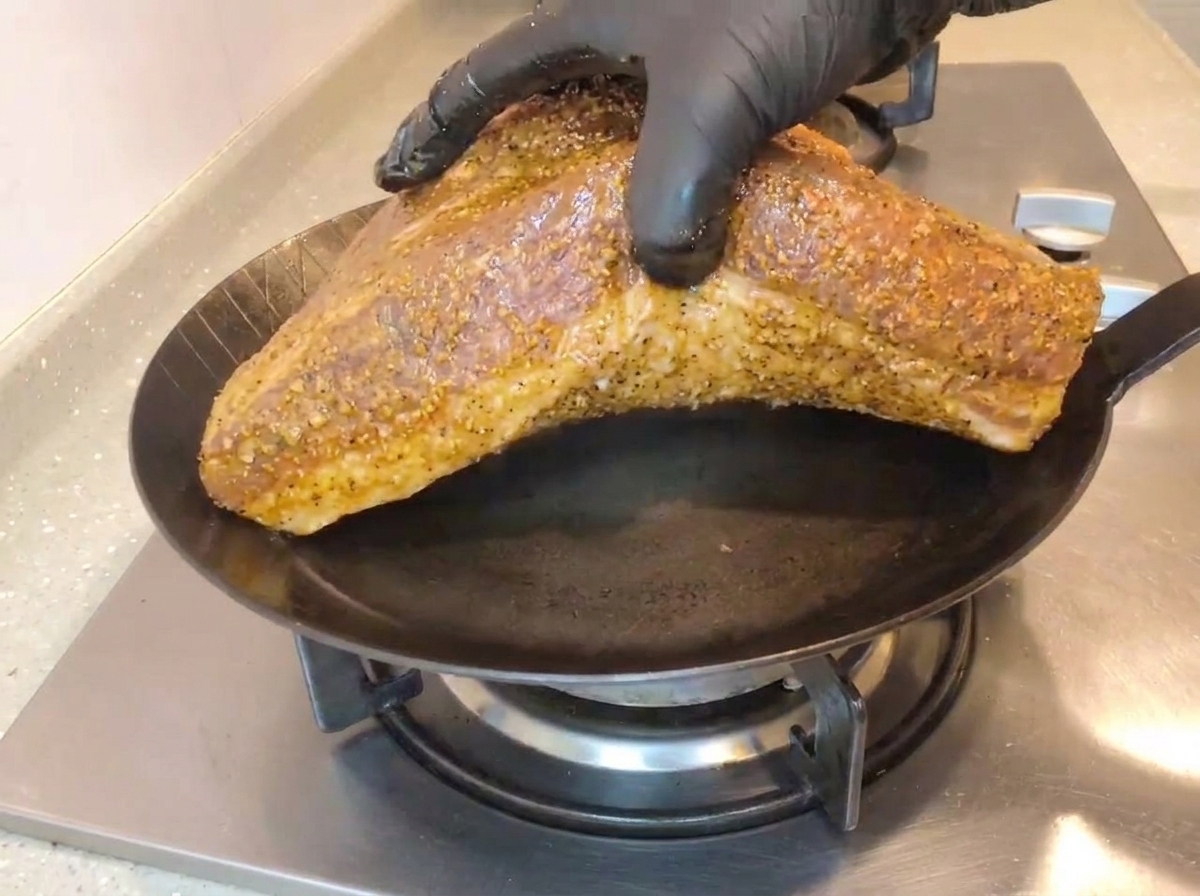 A hand wearing a black glove placing a large, seasoned beef brisket fat-side down into a hot black iron skillet on a gas stovetop.