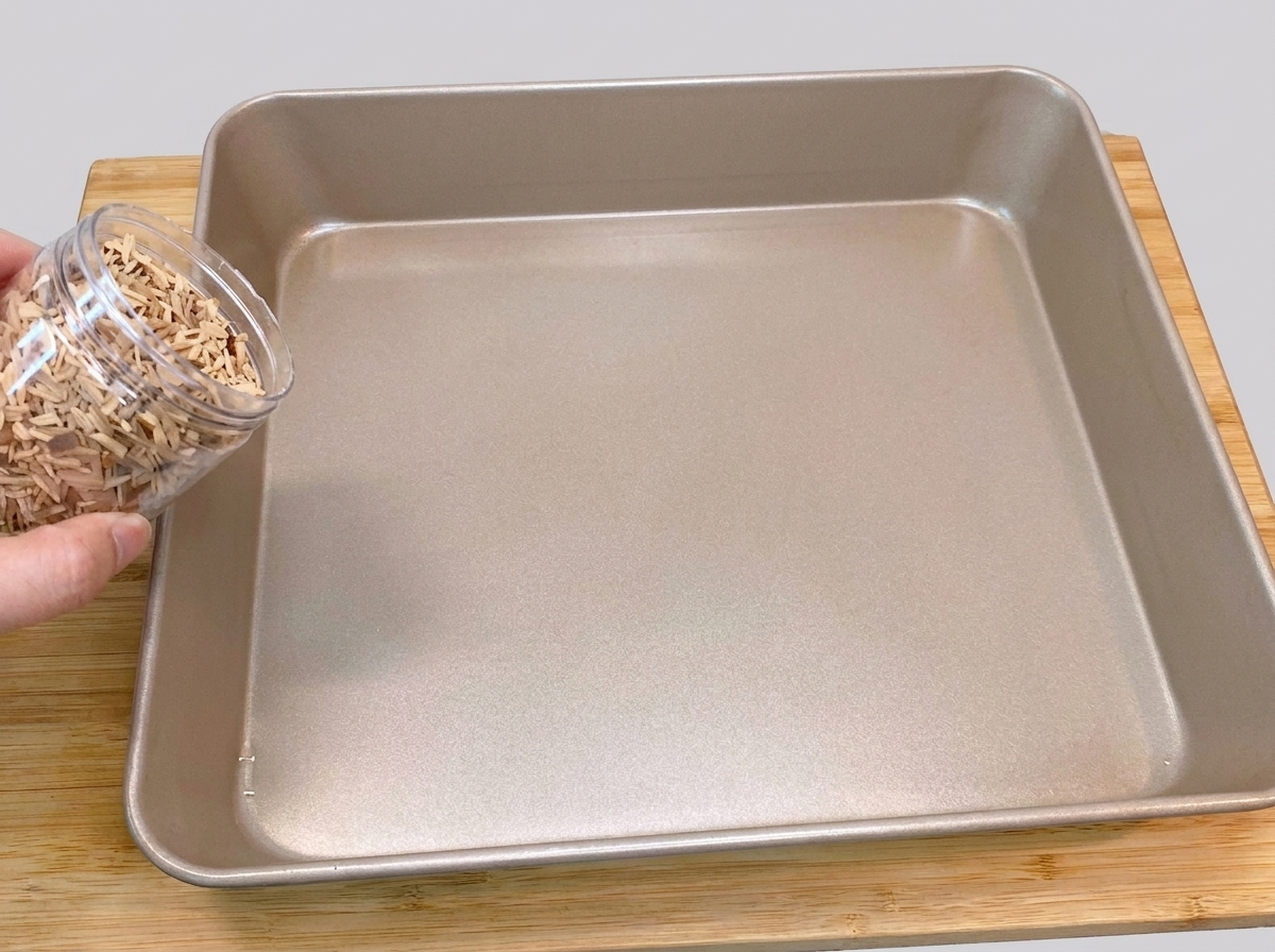A hand pouring peach wood chips out of a plastic container onto the bottom of a square gold-colored baking pan.