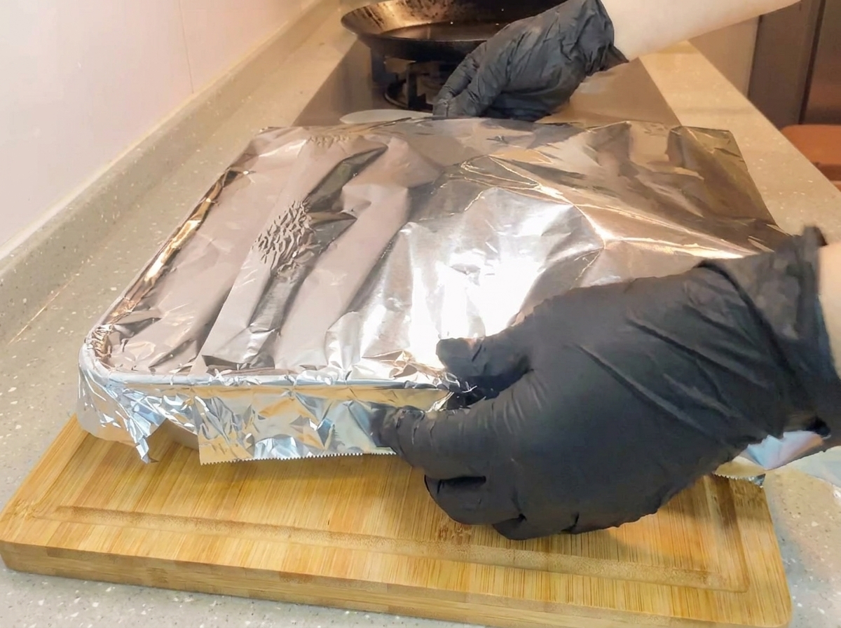 Gloved hands wrapping a sheet of aluminum foil tightly over a square baking pan on a wooden cutting board.