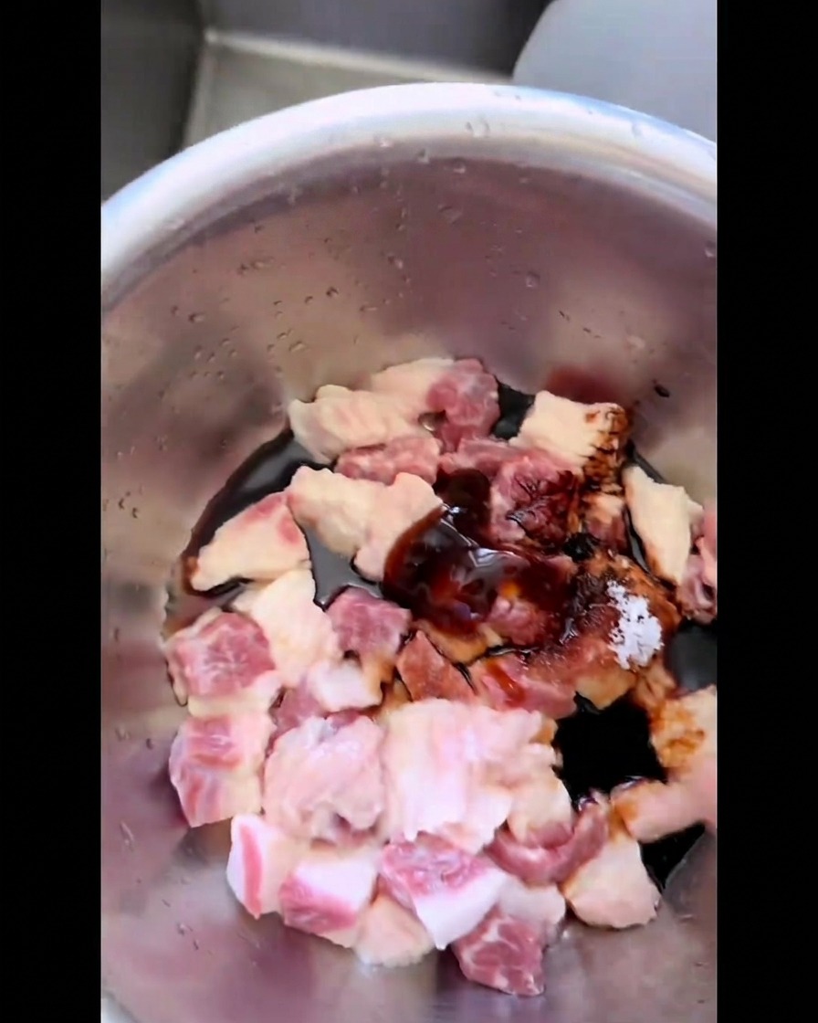 Raw pieces of beef brisket fat inside a stainless steel bowl being combined by hand with savory dark marinade sauces, sugar, and salt.