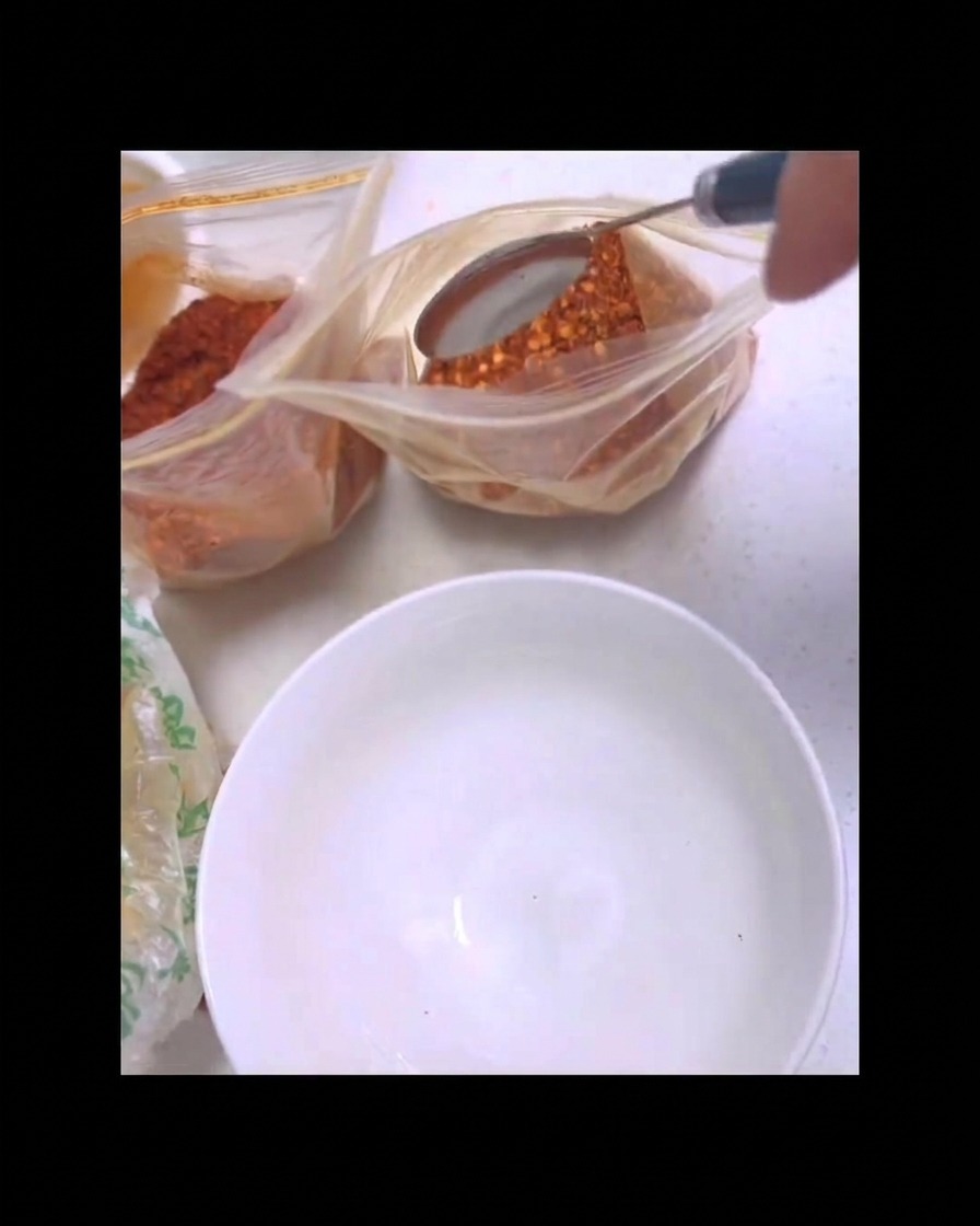 A metal spoon scooping red chili powder out of a clear plastic bag over a clean white bowl.