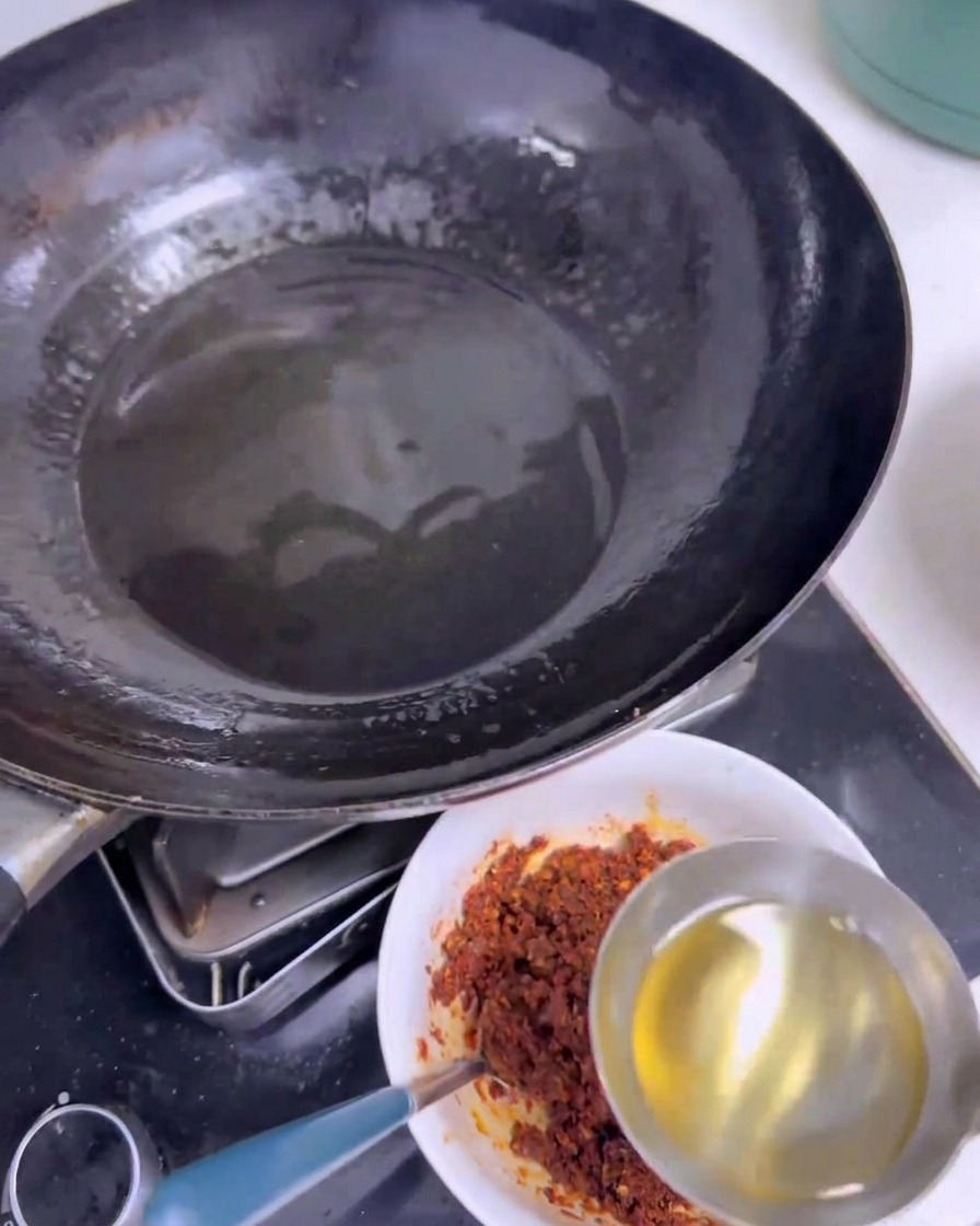 A stainless steel ladle pouring clear hot oil into a white bowl filled with a dark red mixture of chili powder and spices next to a black wok.