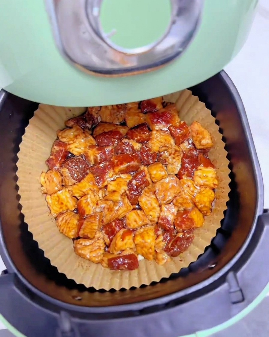 Marinated chunks of beef brisket fat laid out uniformly in a single layer on brown parchment paper inside a green air fryer basket.