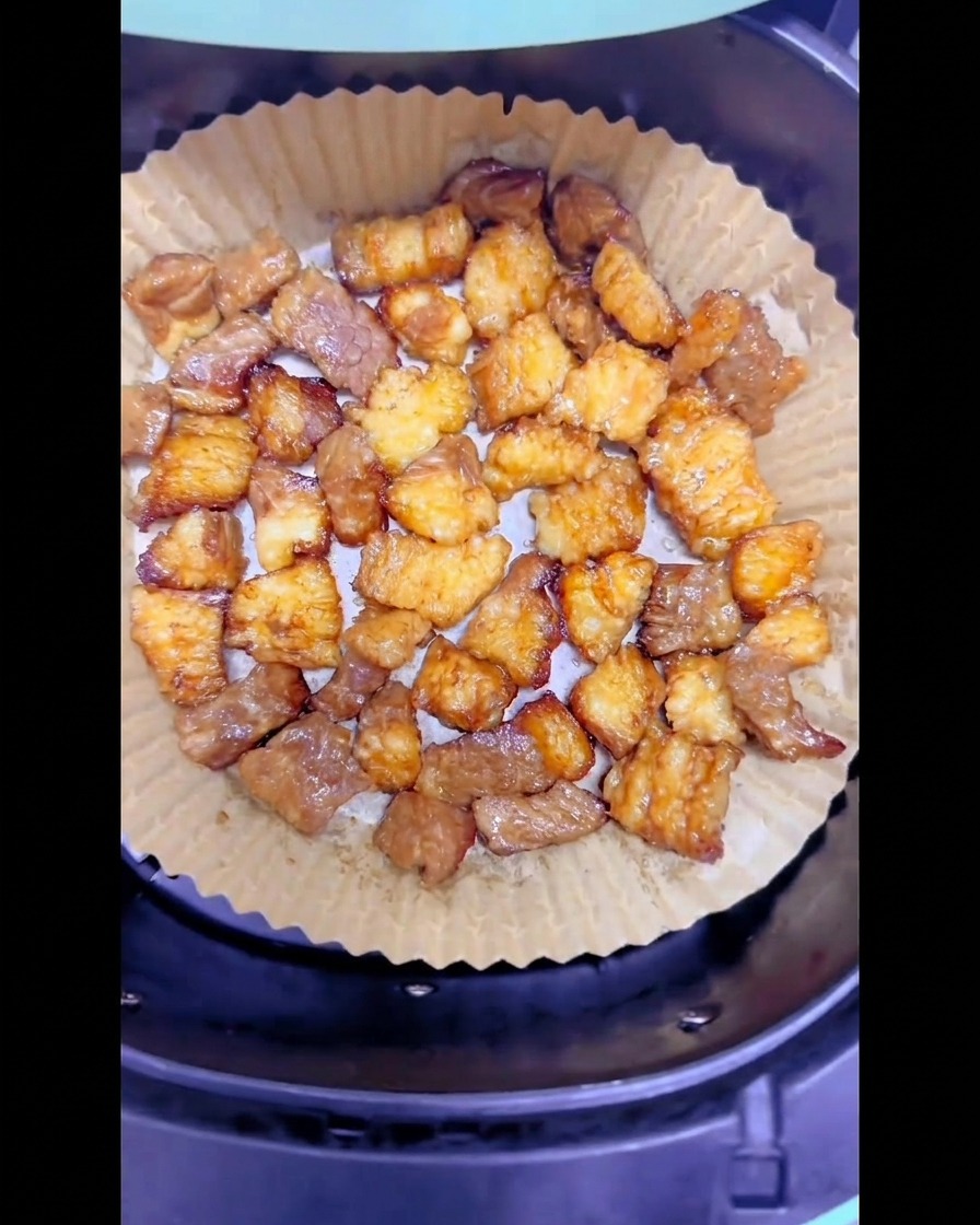 Partially browned pieces of beef brisket fat arranged evenly on a round sheet of parchment paper inside an air fryer basket.