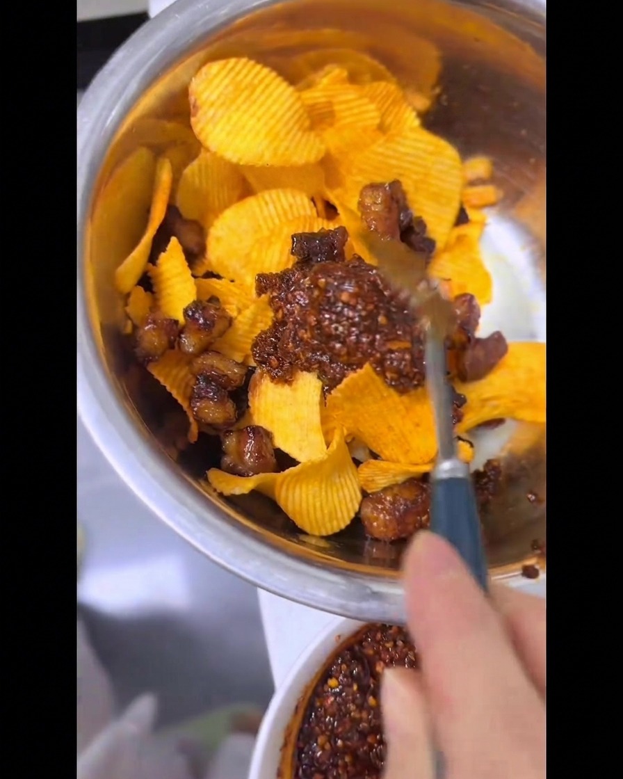 A hand using a spoon to thoroughly mix ridged potato chips, crispy air-fried beef fat, and vibrant red chili oil together in a metal bowl.
