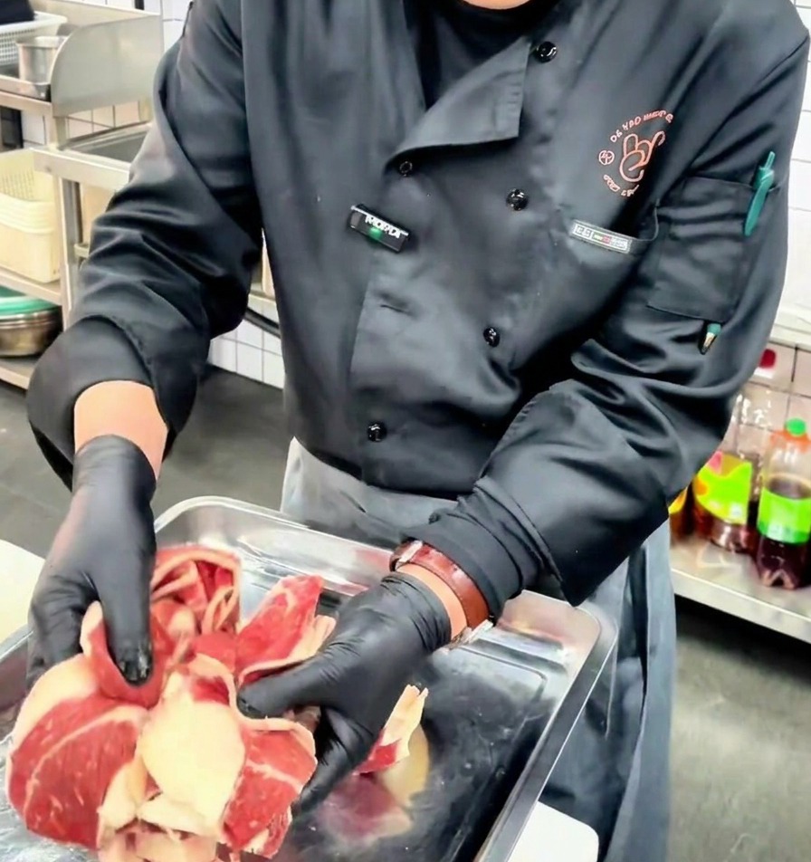 A chef wearing black gloves holding up thin slices of raw beef chest fat with distinct layers of fat and lean meat over a metal tray.