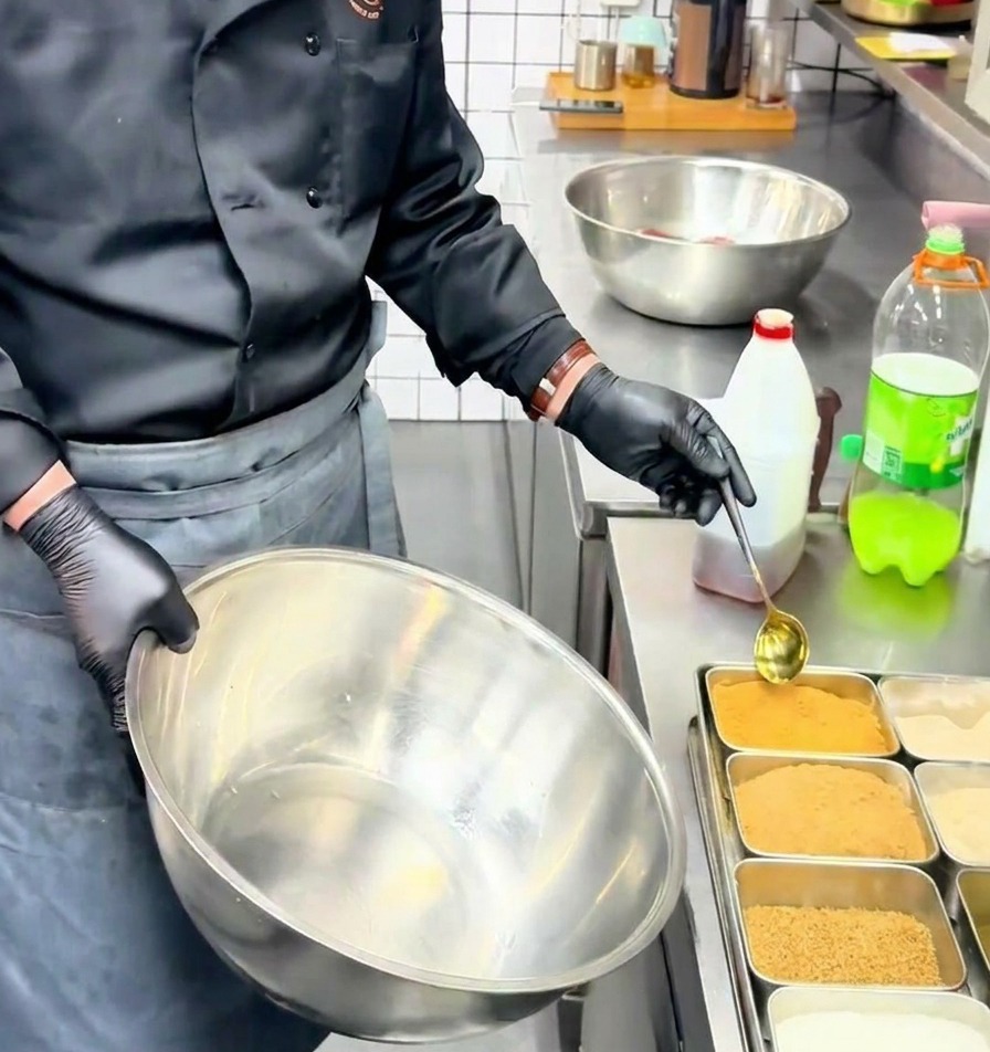 A chef using a golden spoon to scoop yellow beef powder seasoning into a large stainless steel mixing bowl.