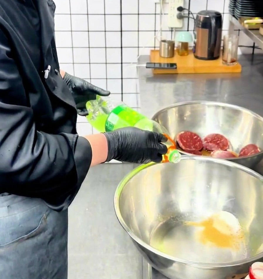 A chef pouring a clear green liquid from a bottle into a stainless steel mixing bowl filled with seasoning powder.