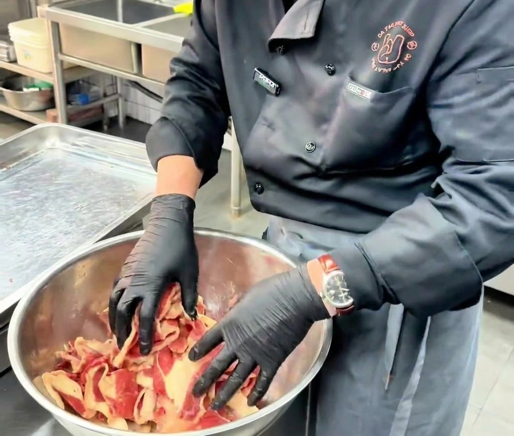 A chef using black gloved hands to mix and massage thinly sliced raw beef with marinade in a large stainless steel bowl.
