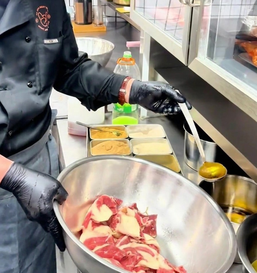 A ladle pouring golden seasoned oil onto marinated beef slices inside a stainless steel mixing bowl.