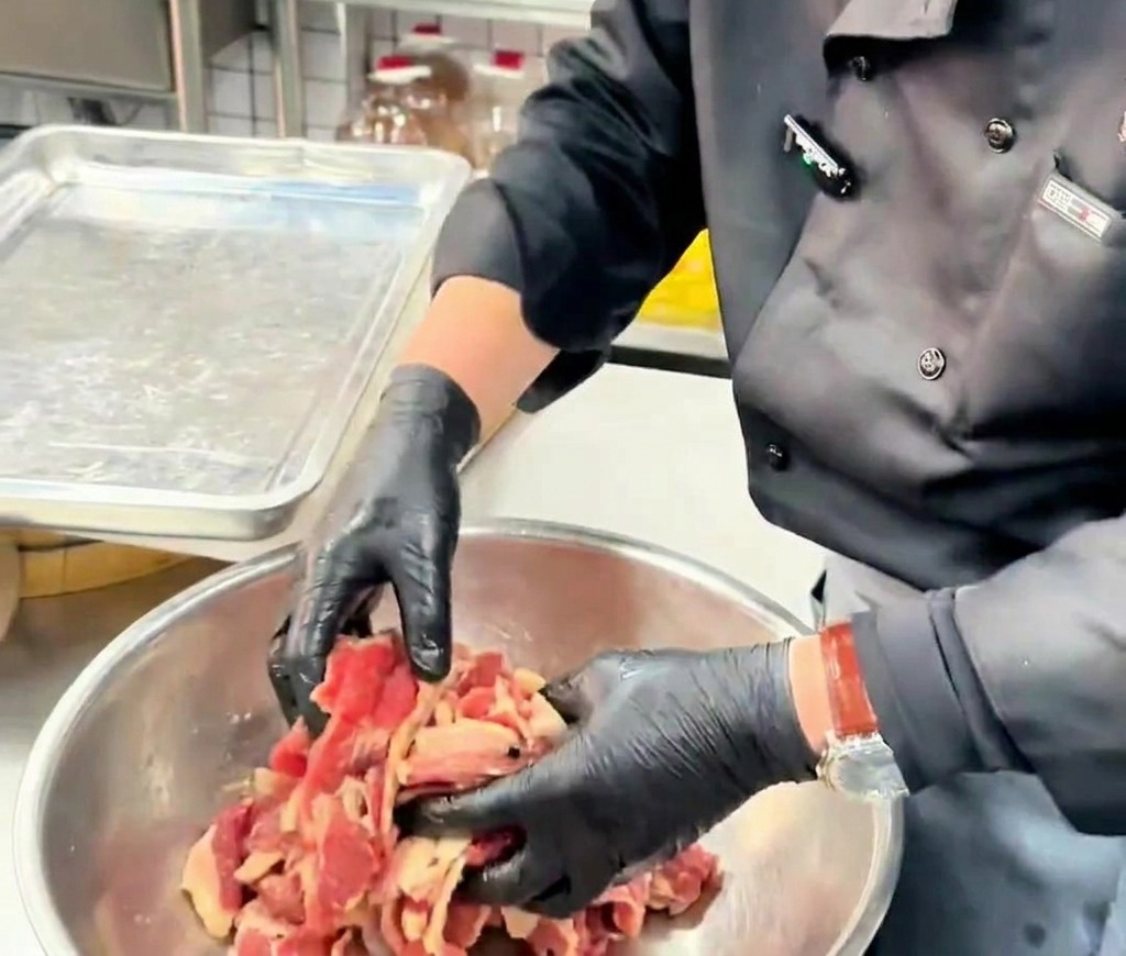Gloved hands tossing and coating thin beef slices with oil inside a metal bowl.