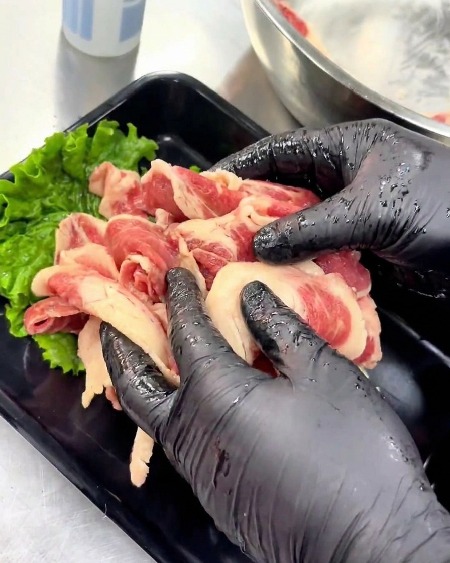 Gloved hands neatly arranging marinated raw beef slices onto a black serving tray decorated with a green lettuce leaf.