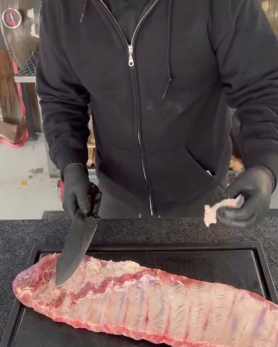 A person wearing black gloves trims excess meat and uneven edges from a raw rack of pork ribs using a large chefs knife on a black cutting board.