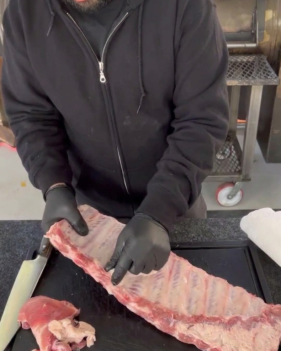 Gloved hands peeling the shiny, translucent membrane away from the back bones of a raw slab of pork ribs on a cutting board.