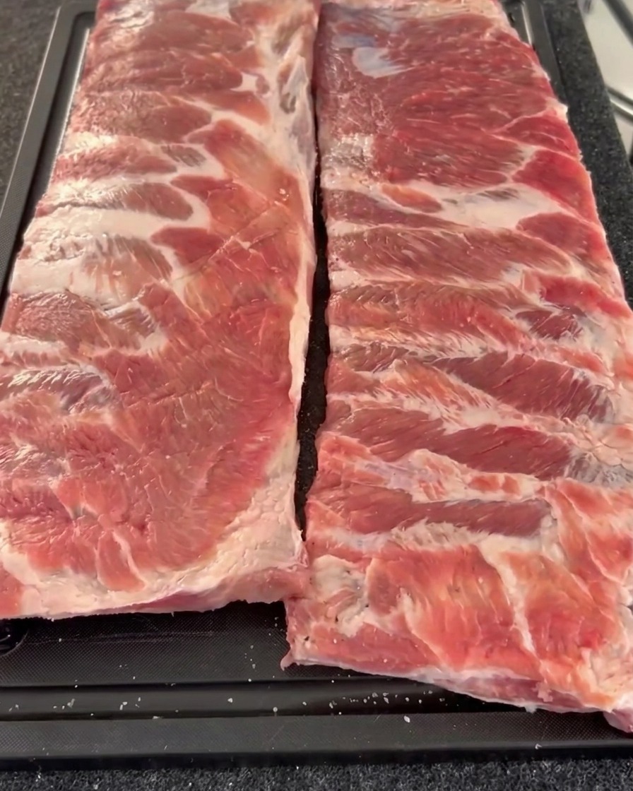 Two raw racks of pork ribs arranged side by side on a cutting board, displaying their seasoned meat side covered in coarse pepper and salt.