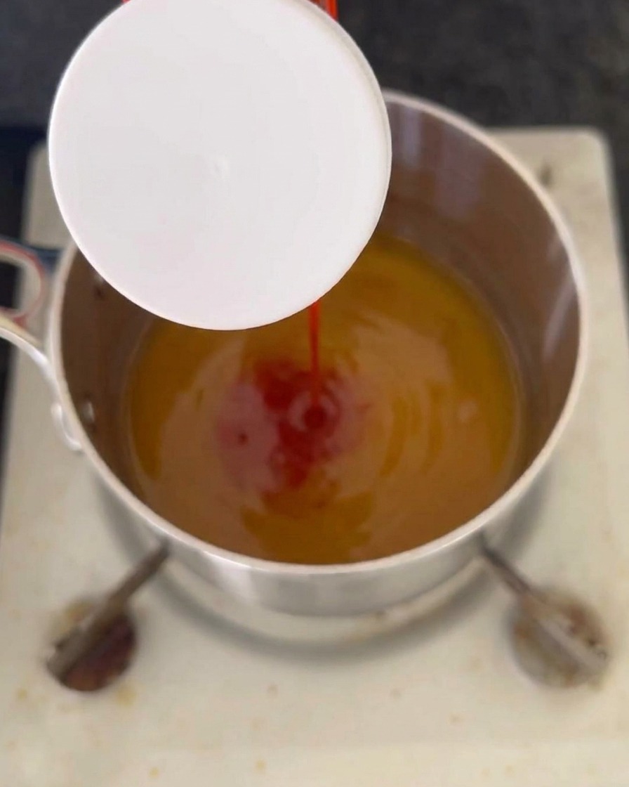 A stream of red ketchup being poured into a bubbling amber liquid mixture inside a stainless steel pot on the stove.
