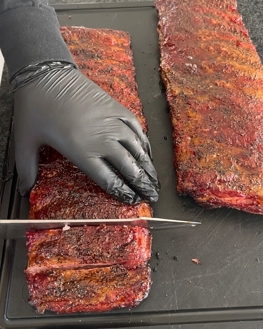 A person wearing a black glove steadying a deeply barked, smoked rib rack while cutting individual ribs with a large knife.