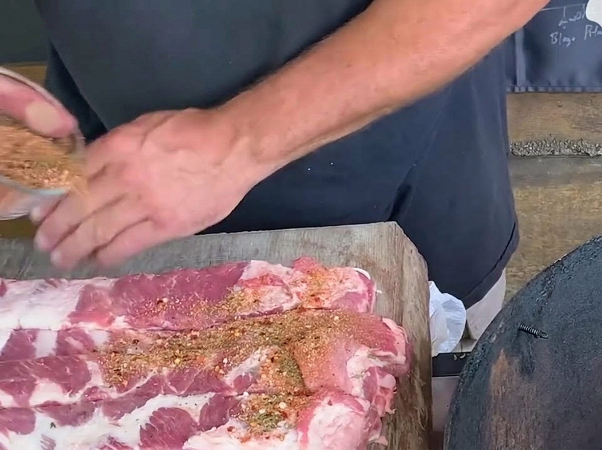 A person sprinkling a spice rub mixture from a glass bowl onto long cut strips of raw pork ribs on a rustic wooden board.