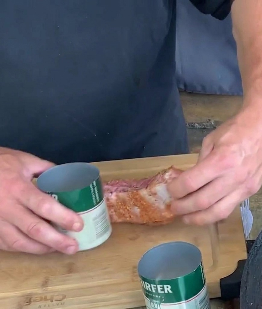 Hands shaping a seasoned pork rib strip by wrapping it around an open green and white aluminum can on a cutting board.